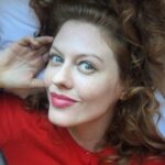 A woman with curly hair wearing a red top, smiling at the camera